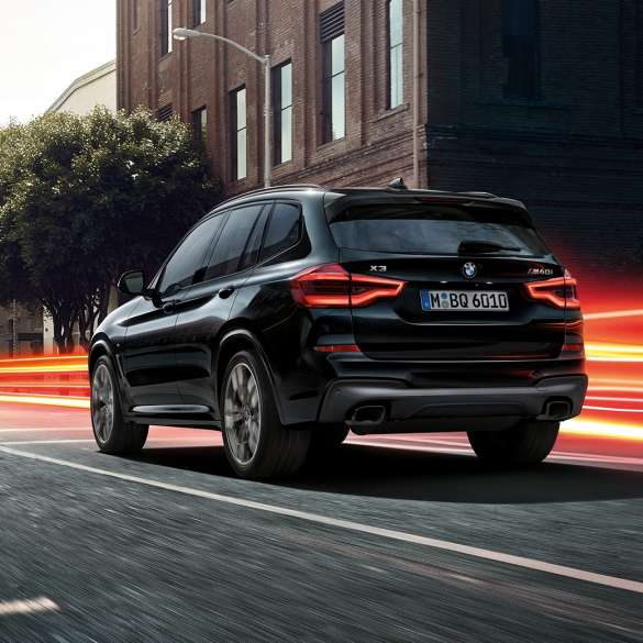 BMW X3 M40i, vue de trois quarts arrière. Extérieur de la BMW X3 M40i en Saphierschwarz metallic, en mouvement, vue de trois quarts arrière devant un paysage urbain avec bandes lumineuses.