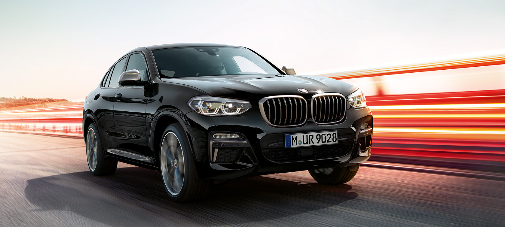 BMW X4 M40i et BMW X4 M40d, vue de trois quarts avant gauche. Extérieur des BMW X4 M40i et BMW X4 M40d en Saphierschwarz metallic, en mouvement, vue de trois quarts avant gauche avec bandes lumineuses.