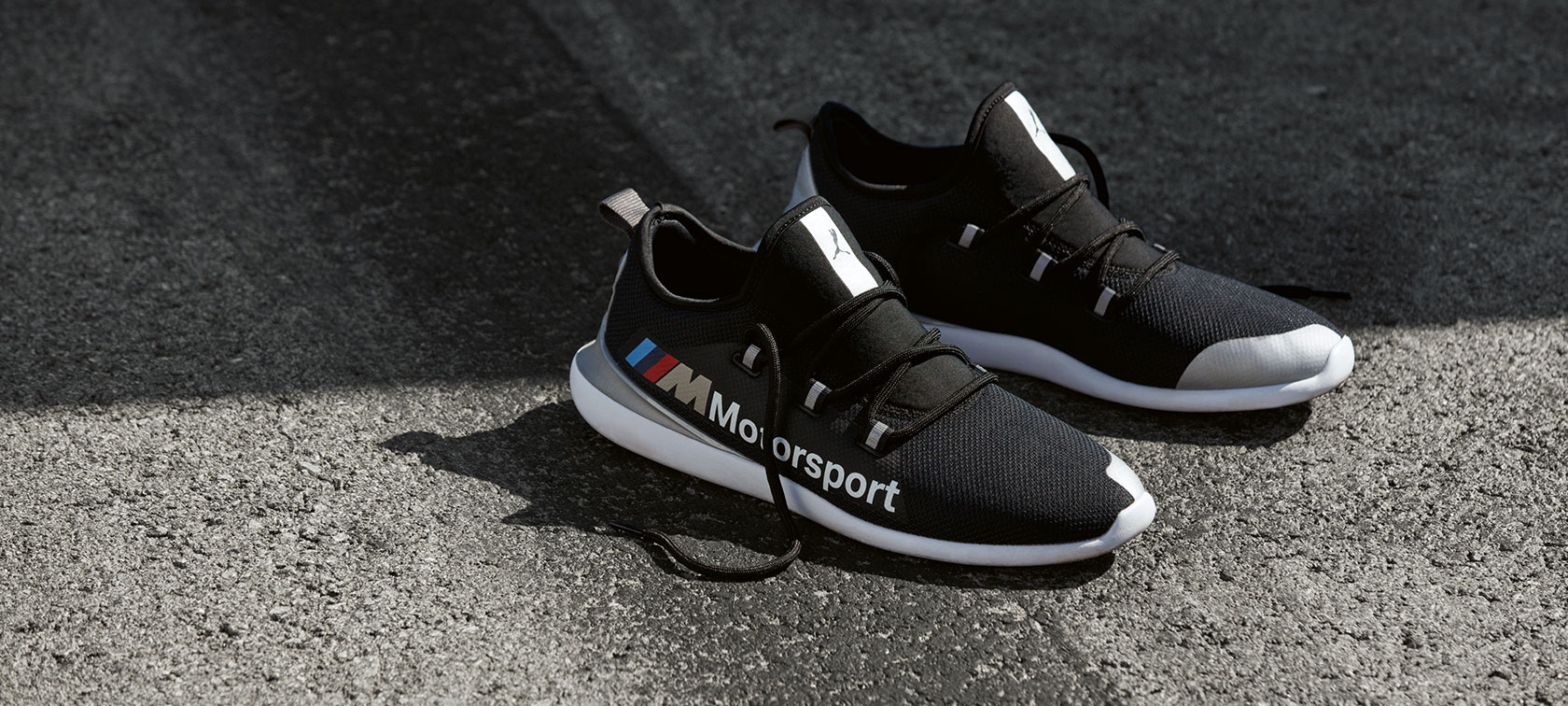 Chaussures BMW M Motorsport PUMA EVO CAT. Une paire de chaussures BMW M Motorsport PUMA EVO CAT de la Collection BMW M Motorsport est présentée sur une surface de tarmac.
