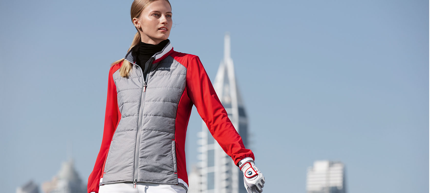 Veste femme BMW Golfsport. La jeune femme de la photo présente des articles premium de la Collection BMW Golfsport contre un décor de grande ville.