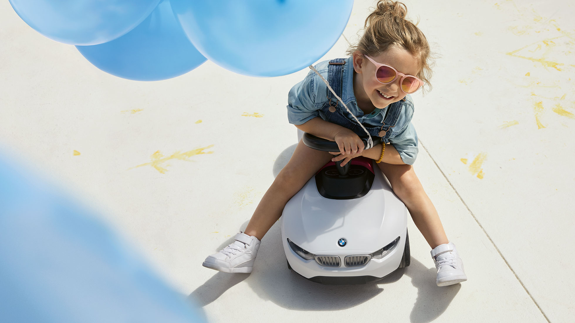 Collection BMW Kids. Une fillette joyeuse avec des lunettes de soleil roses et un ballon bleu est assise sur une BMW Baby Racer.