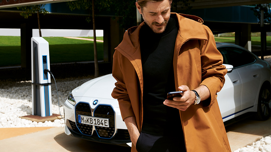 BMW Serenity durées et échéances des véhicules électriques BMW Serenity, un homme regarde son smartphone devant une BMW i4 à une borne de recharge